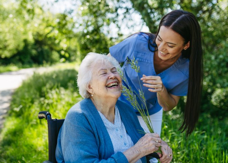An elderly lady in a wheelchair outdoors, bright sunshine, standing beside her is her live-in carer
